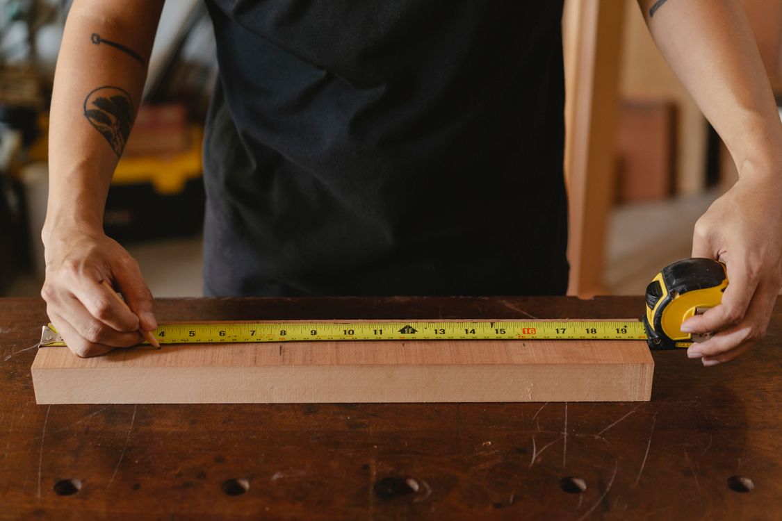 Woodworker measuring a wooden plank with a tape measure at a workshop bench.